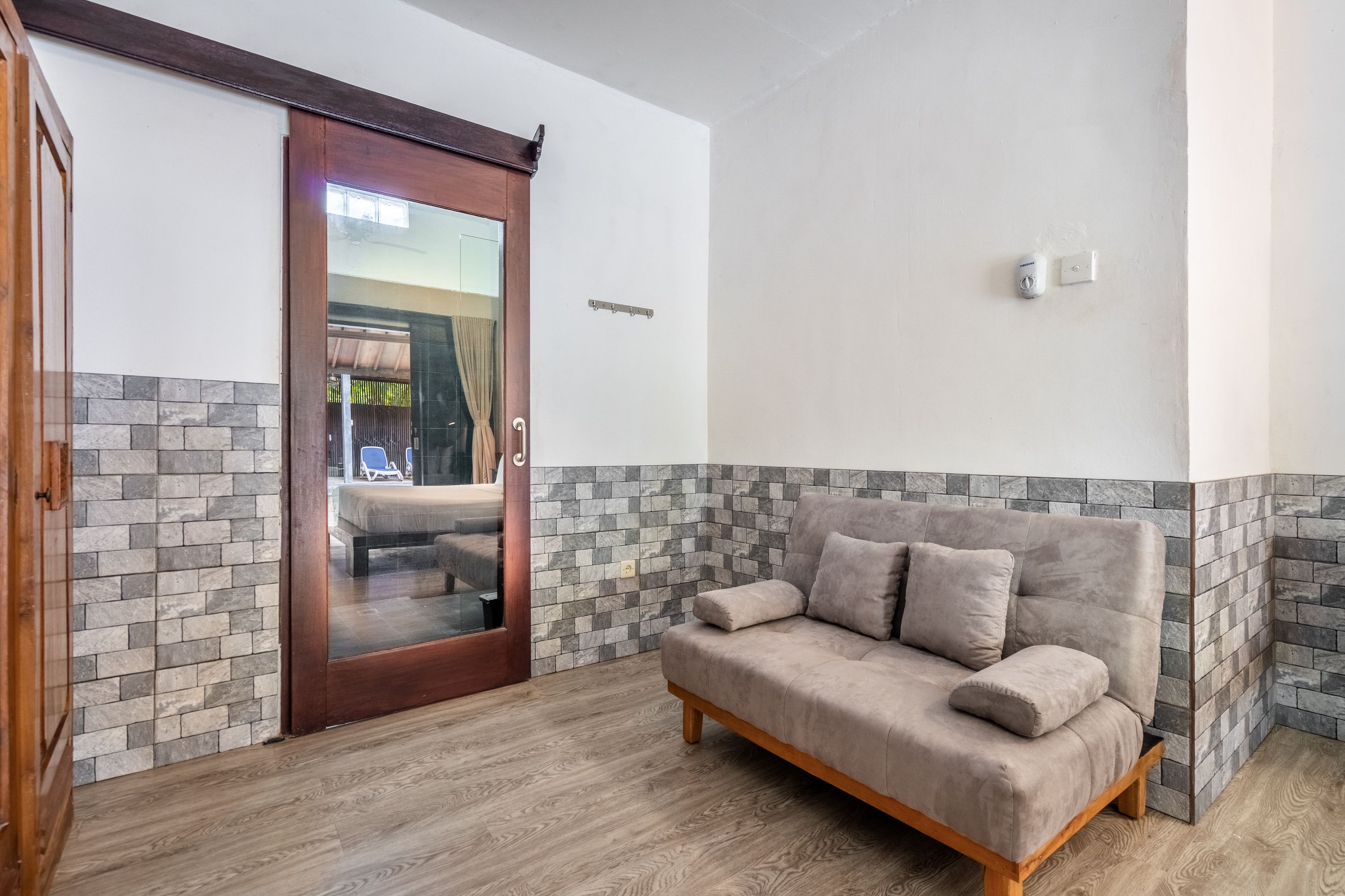 Una habitación con puerta de madera, pared con baldosas de piedra, sofá gris y suelo de madera.