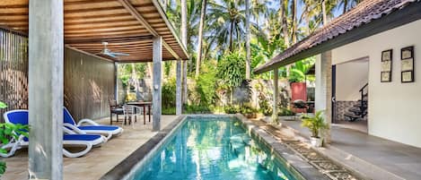 A rectangular swimming pool with clear blue water, surrounded by a tiled deck. There are lounge chairs and a covered seating area with a table and chairs. The pool is adjacent to a building with a tiled roof and a wall with framed artwork.