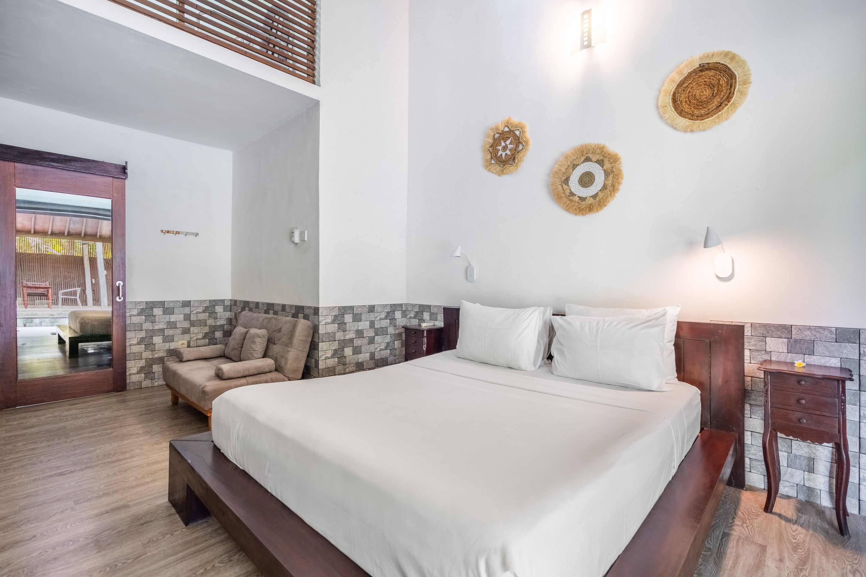 A hotel room with a large bed, a wooden bench, a stone wall, and a view of the outdoor area.