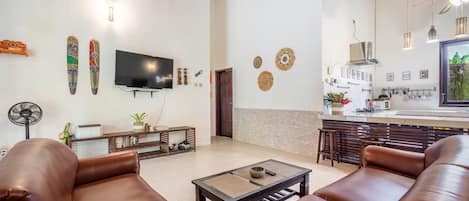 A modern living room with leather sofas, a flat-screen TV, and a dining area with a kitchen in the background.