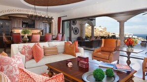 A spacious living area with a white sofa, a leather armchair, and a wooden coffee table. There is a kitchen in the background with modern cabinetry and a large flat-screen TV. The room features a large window with a view of the ocean and a balcony area with outdoor seating. - Fundadores 121 - Puerto Los Cabos (San Jose Del Cabo)