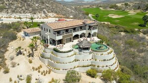 A large, modern two-story building with a curved, multi-level pool area and a golf course in the background. - Fundadores 121 - Puerto Los Cabos (San Jose Del Cabo)