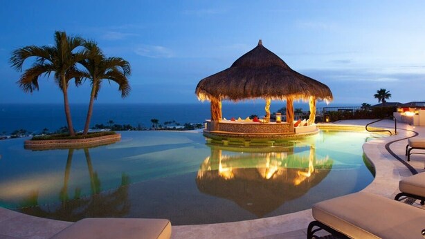 A infinity pool with a thatched gazebo and lounge chairs, palm trees, and a view of the ocean. - Fundadores 121 - Puerto Los Cabos (San Jose Del Cabo)