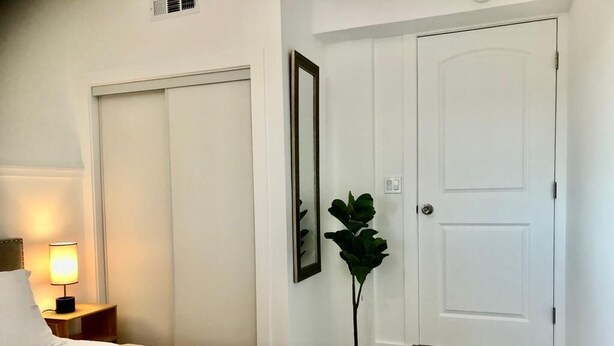 A hotel room with a bed, a nightstand with a lamp, a potted plant, and a door. - Private 1BR Suite w Full Kitchen & Laundry (Hayward)