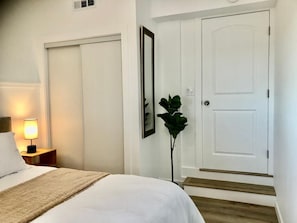 A hotel room with a bed, a nightstand with a lamp, a potted plant, and a door.