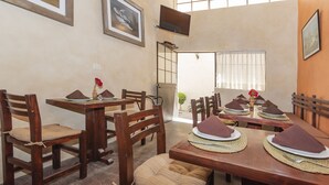 Un comedor con mesas y sillas de madera, algunas con cojines. Hay individuales y un televisor montado en la pared.