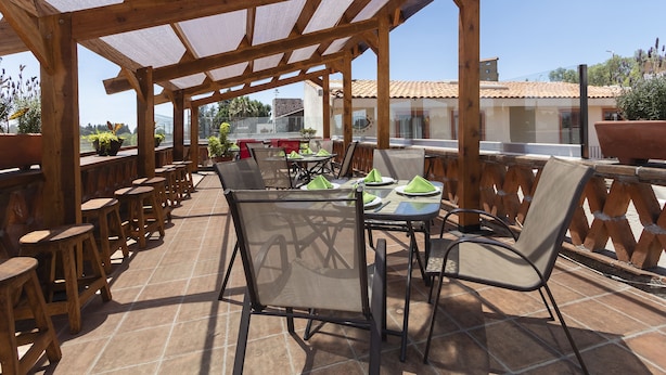 Un patio al aire libre con mesas y sillas de madera, una zona de estar cubierta y vistas a edificios al fondo.