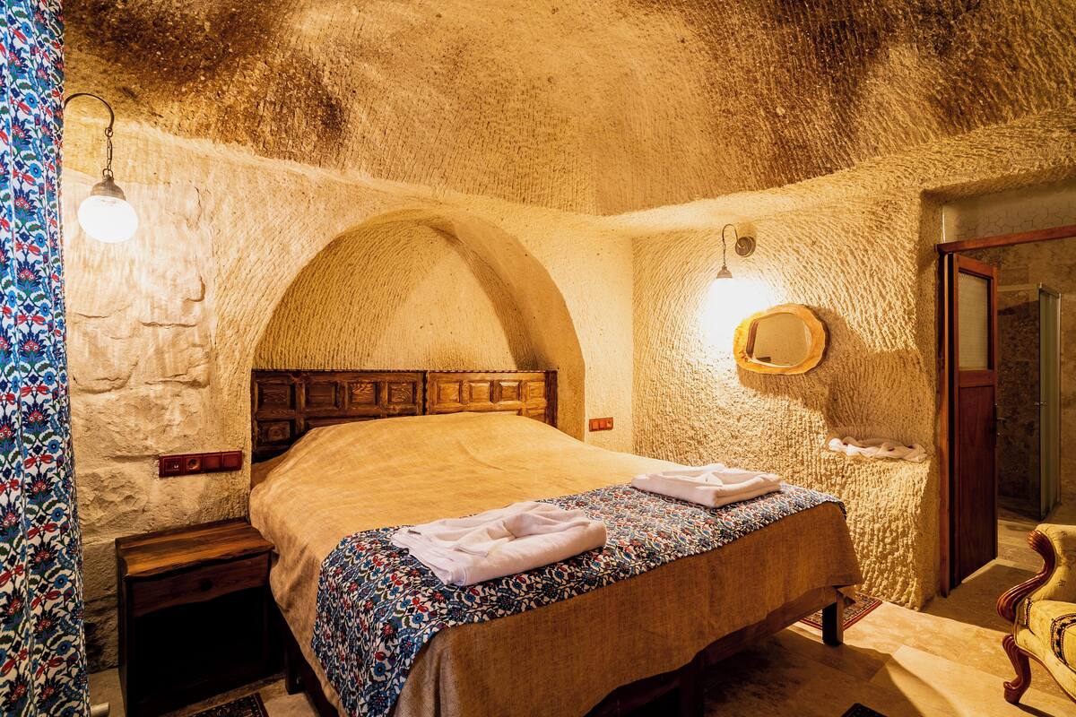 A bedroom with a stone wall, a wooden bed, a nightstand, a mirror, and a patterned bedspread.