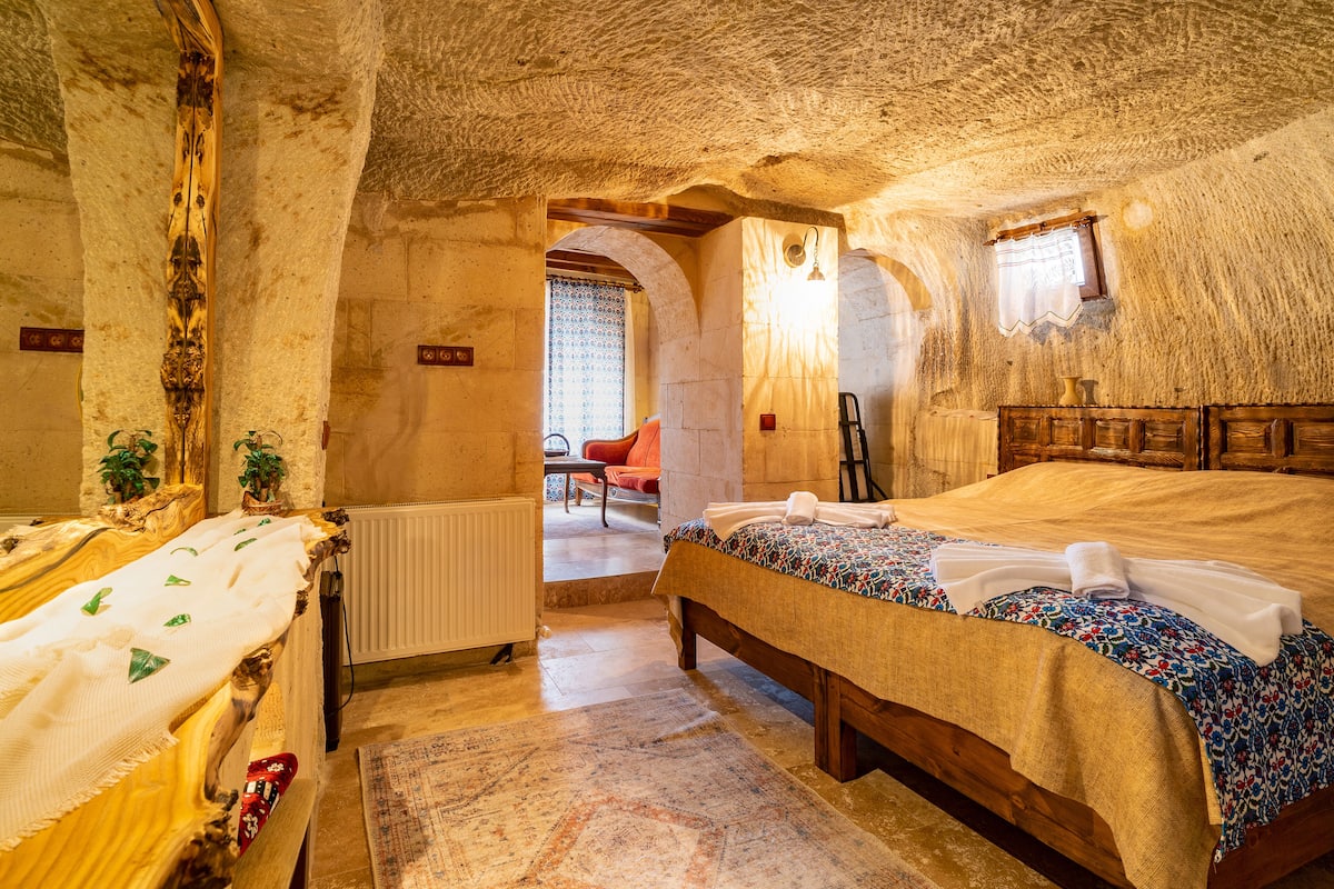 A cozy room with a wooden bed, a small table, and a chair. There is a window with curtains and a view of the outside.