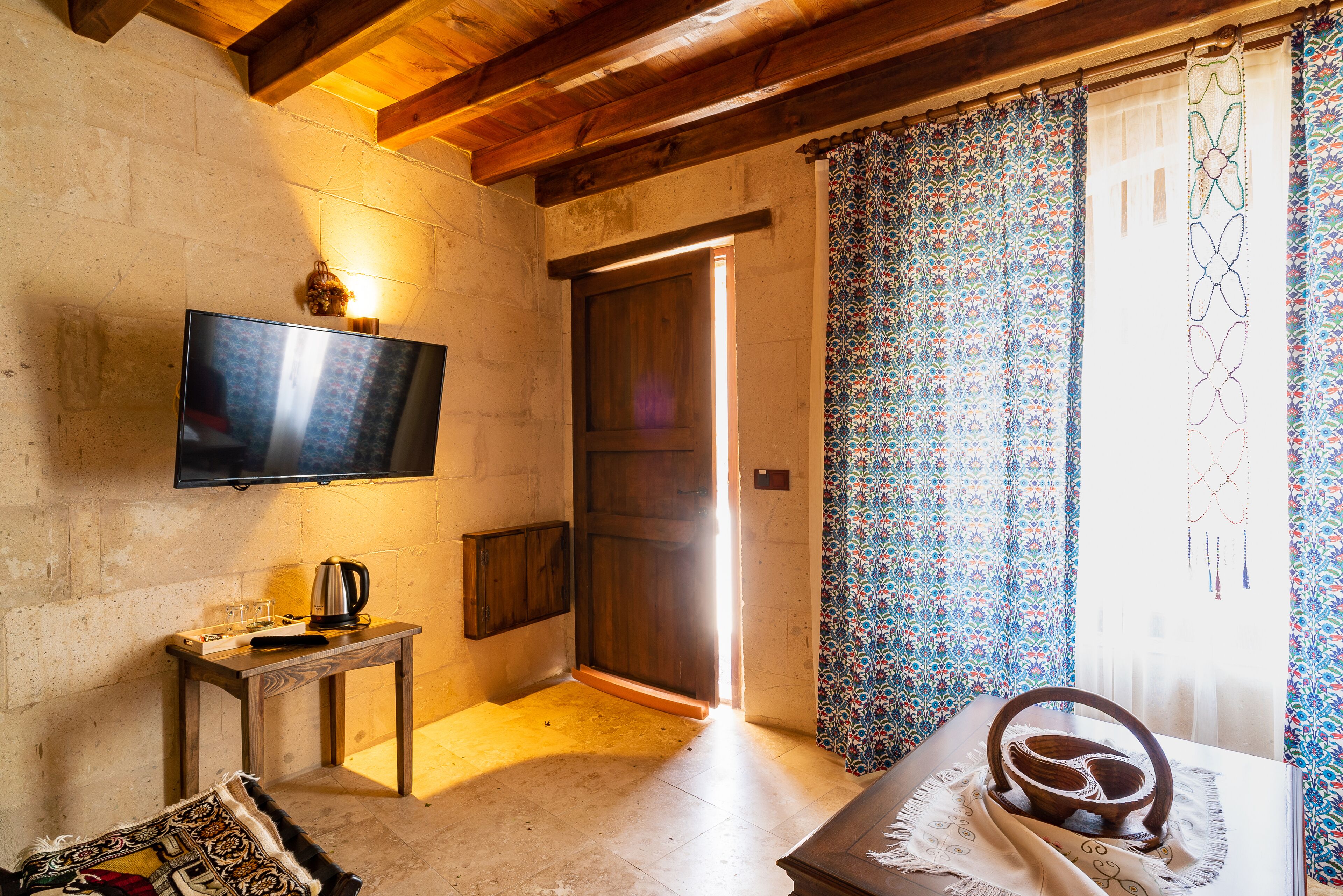 Quarto com televisão na parede, mesa de madeira com chaleira, cortina estampada e porta que dá para o exterior.
