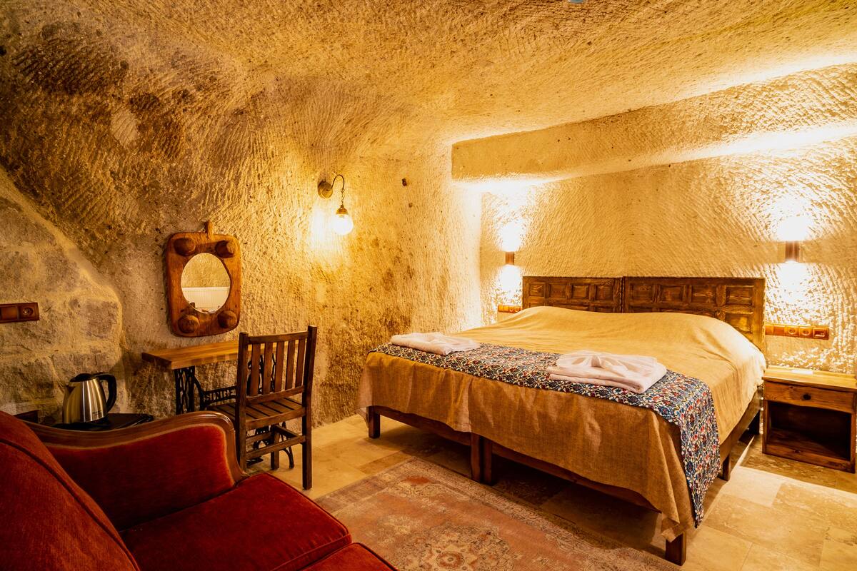 A rustic bedroom with a stone wall, a wooden bed, a red chair, and a wooden nightstand with a lamp.
