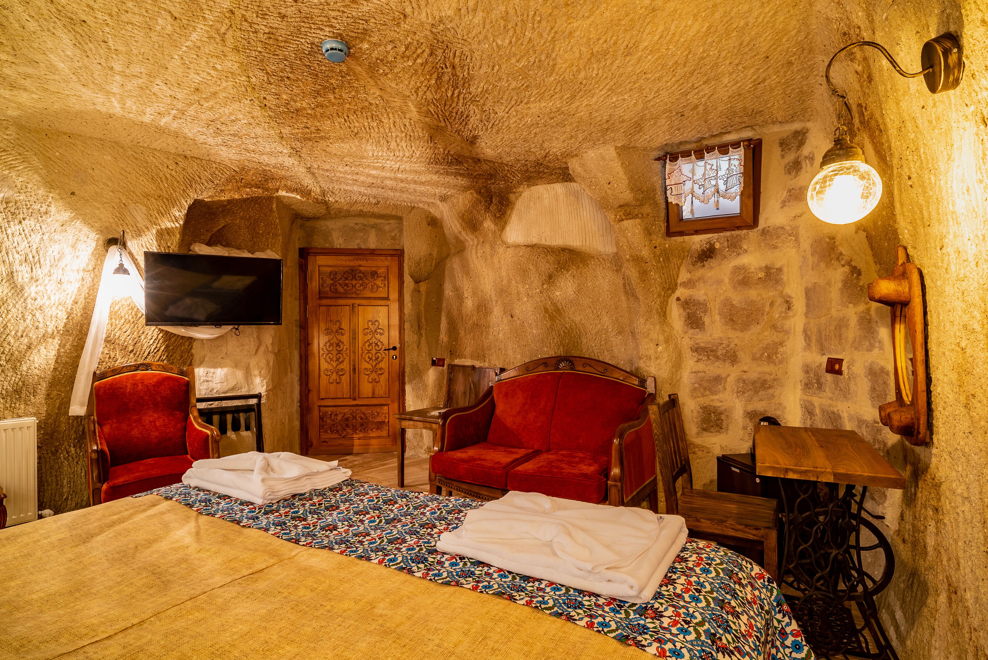 A room with a bed, two red chairs, a wooden cabinet, a television, and a wall-mounted lamp.
