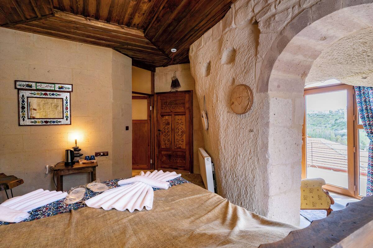 A rustic room with a bed, a wooden desk, a wall-mounted lamp, and a view of the outdoors.