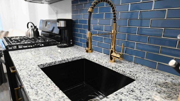 A kitchen with a granite countertop, a stainless steel sink, a gold faucet, and a blue-tiled backsplash.