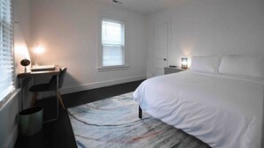A bedroom with a bed, a desk, and a chair. There is a window with blinds, a door, and a nightstand with a lamp.