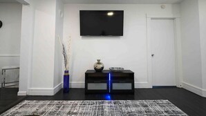 A modern living room with a flat-screen TV mounted on the wall, a dark wooden entertainment unit with blue LED lighting, and a patterned rug on the floor.