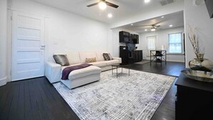 A spacious living area with a white sofa, a coffee table, and a patterned rug. There is a kitchen area with black cabinets and a dining table. The room has a ceiling fan and recessed lighting.