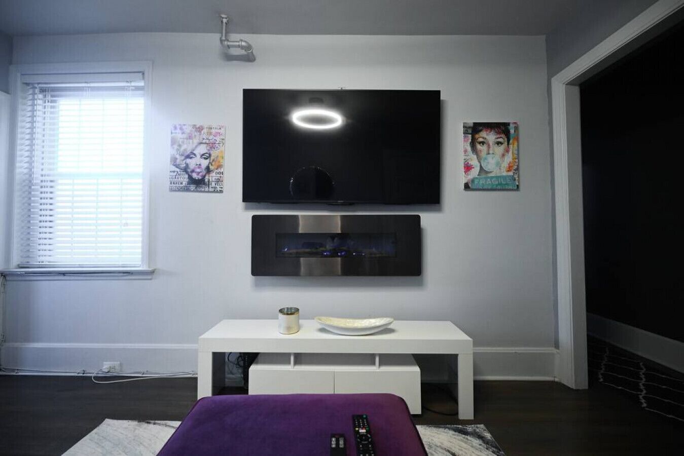 A modern living room with a flat-screen TV mounted above a white console table, a window with blinds, and artwork on the walls.