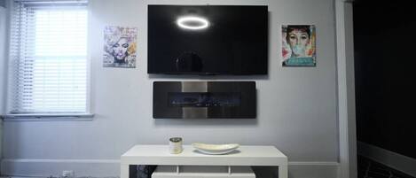 A modern living room with a flat-screen TV mounted above a white console table, a window with blinds, and artwork on the walls.