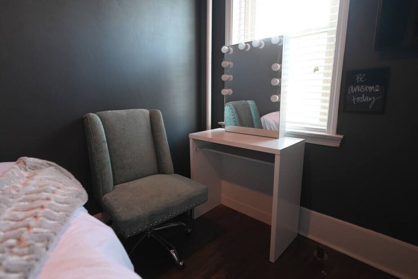 A bedroom with a bed, a chair, a vanity with a mirror, and a wall with a motivational quote.