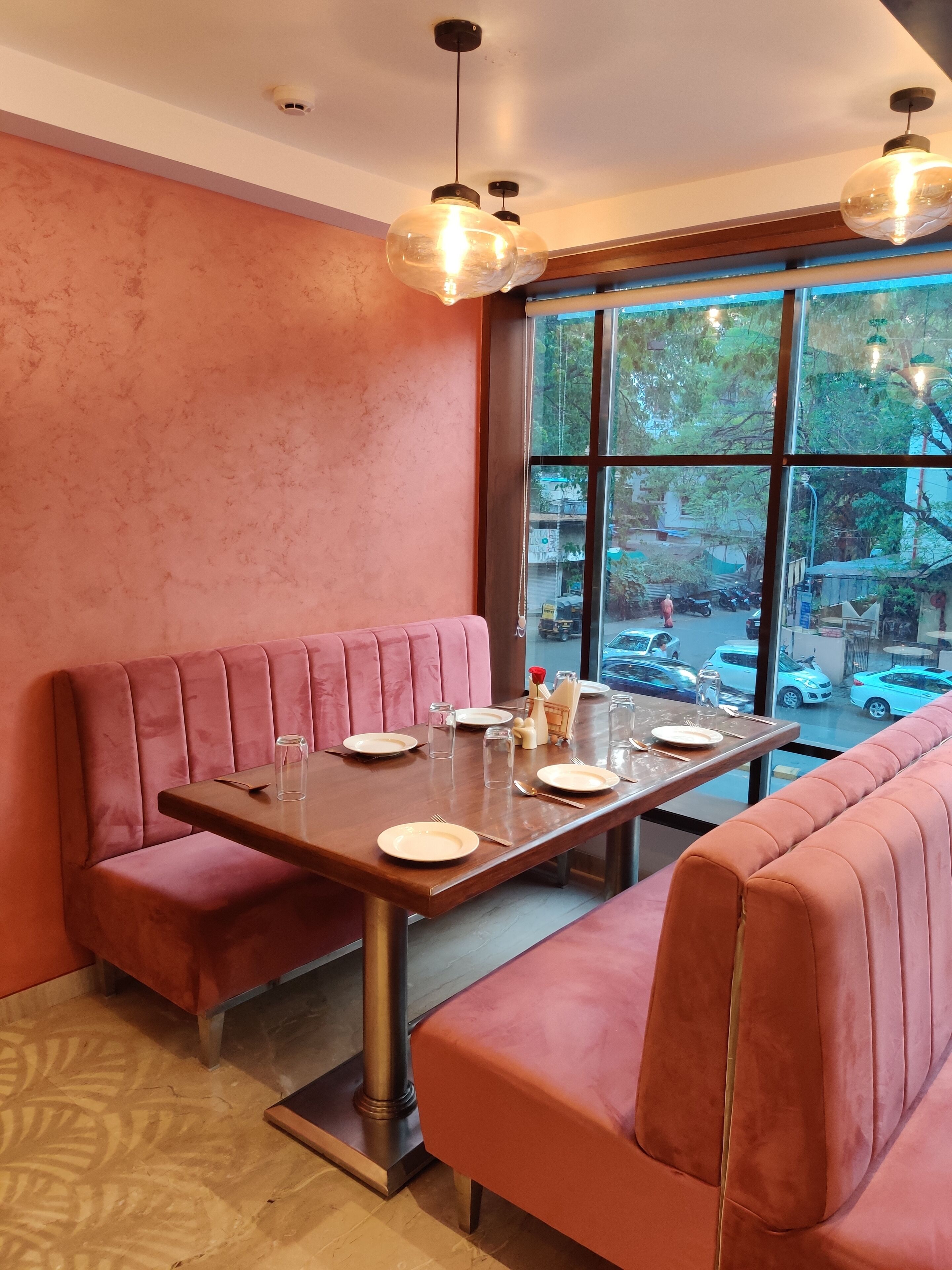 A restaurant with a pink booth seating area, a wooden table set with plates and cutlery, and a view of the street through large windows.