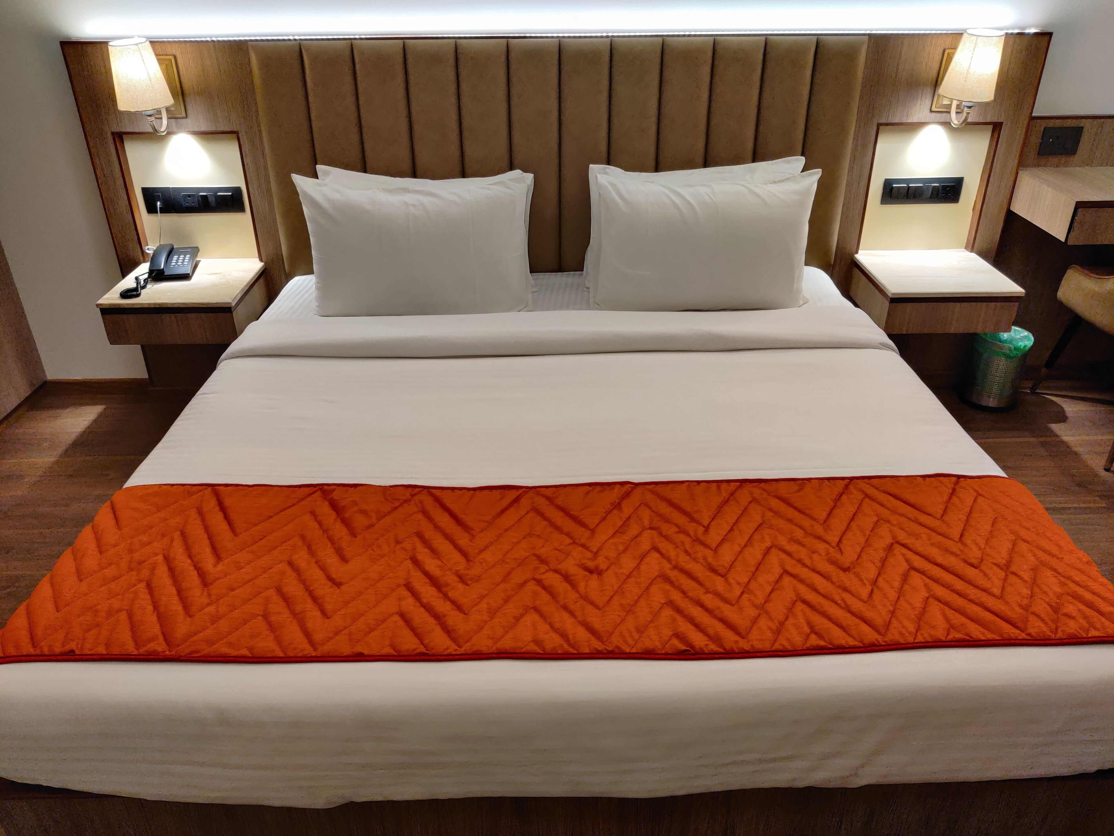 A hotel room with a large bed, two white pillows, a wooden headboard, bedside lamps, a telephone, and a trash bin.