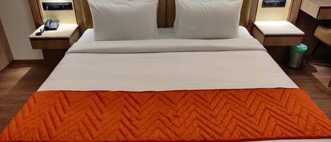 A hotel room with a large bed, two white pillows, a wooden headboard, bedside lamps, a telephone, and a trash bin.