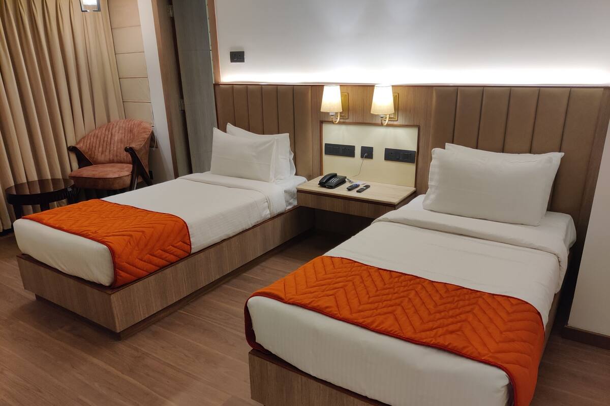 Two single beds with orange bedding, a wooden headboard, a bedside table with a phone and remote controls, and a chair.