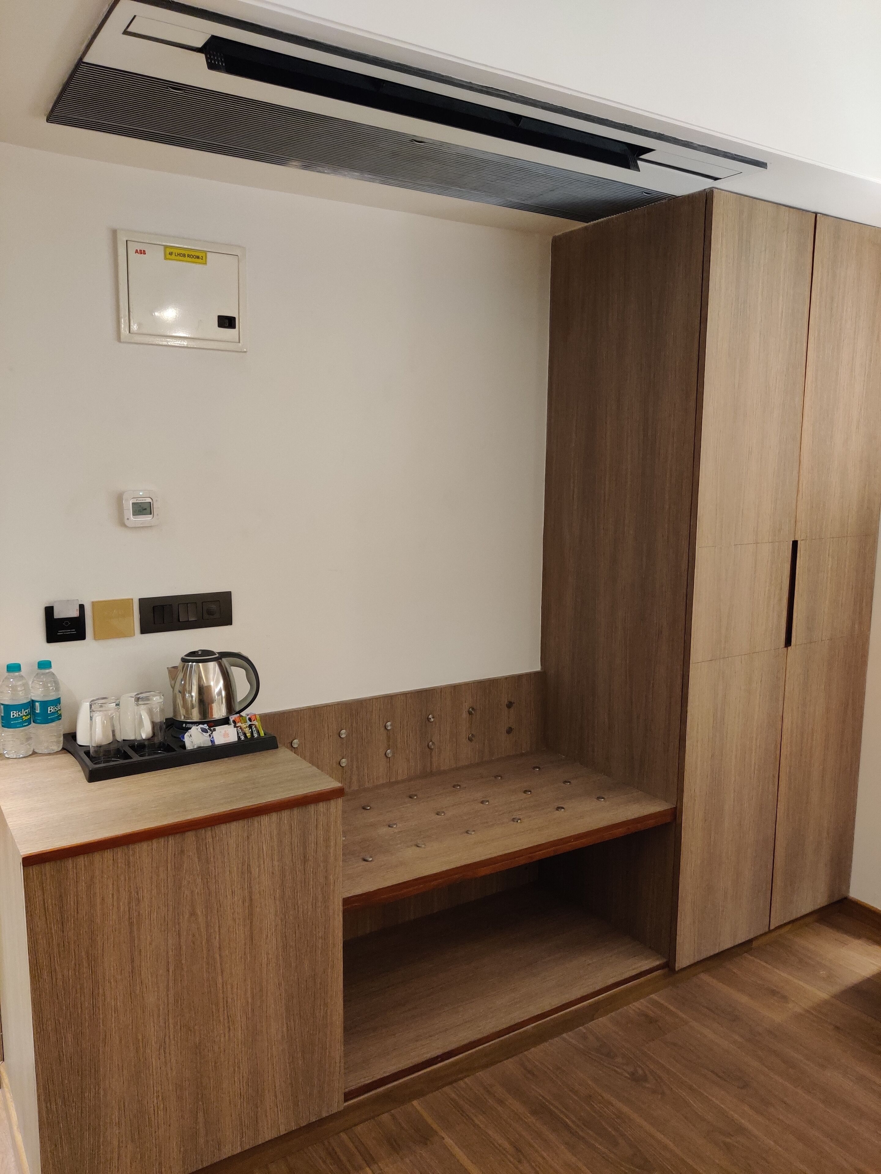 A hotel room with a wooden wardrobe, a bench, a coffee station, and a wall-mounted air conditioning unit.