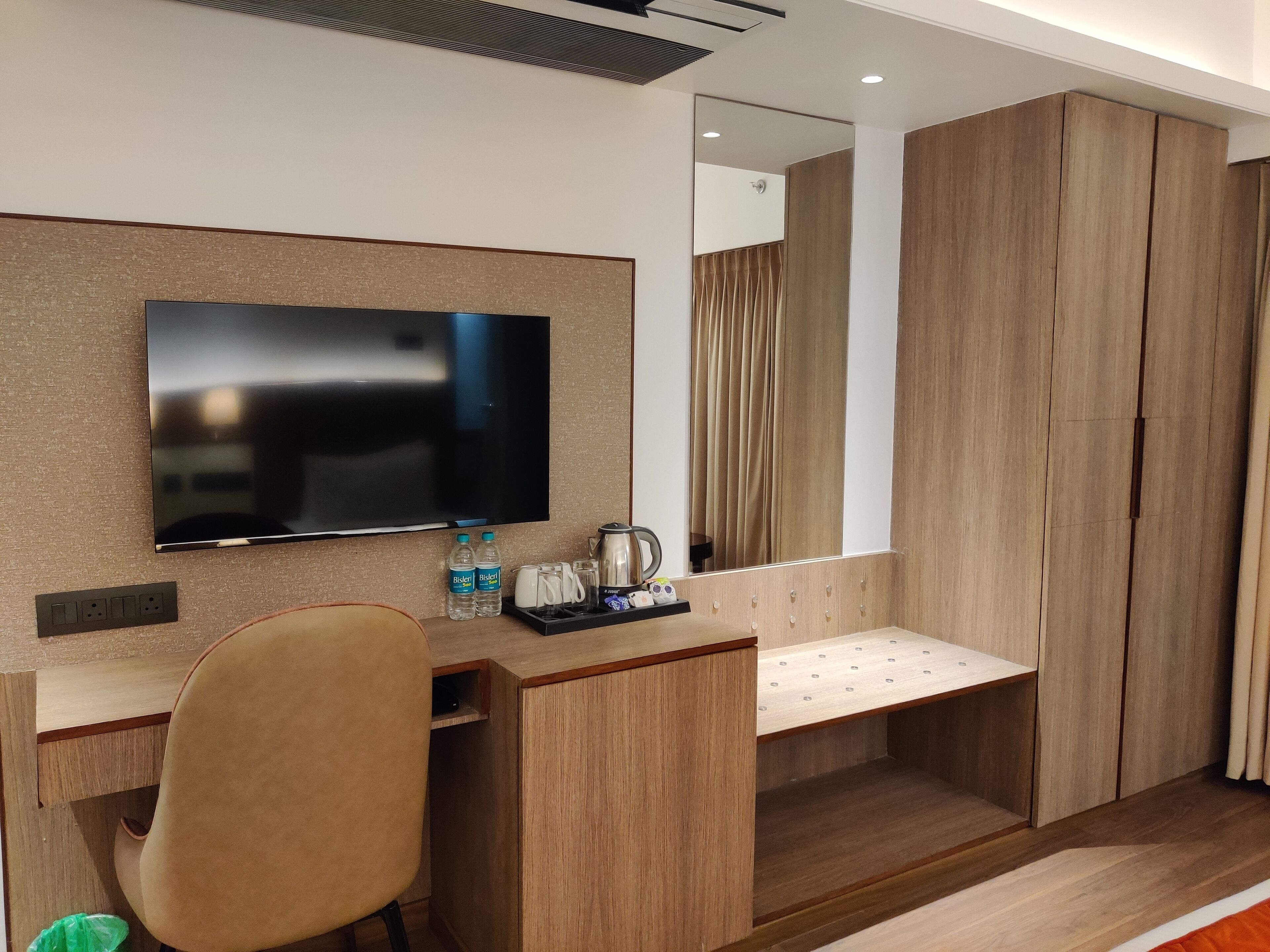 A modern hotel room with a wooden desk, a flat-screen TV, a mirror, and a wardrobe.