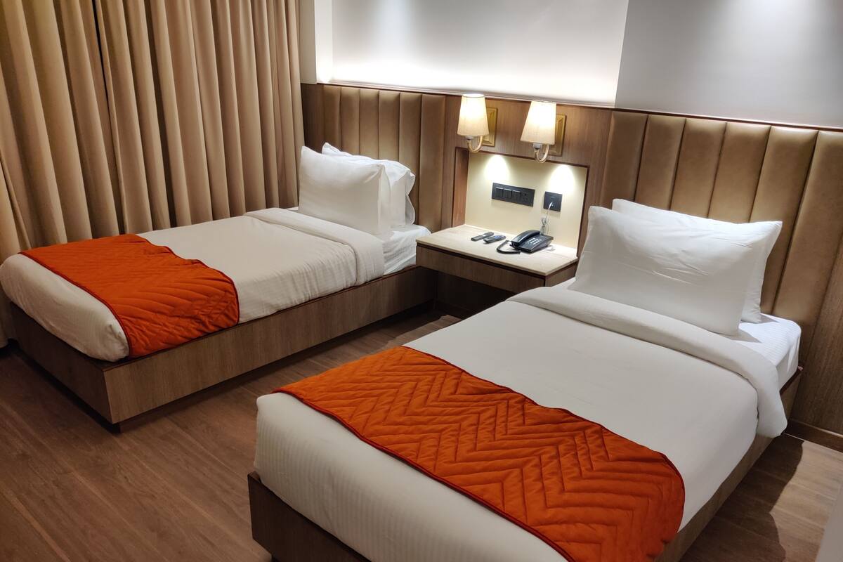 A hotel room with two beds, each with an orange quilted blanket, a wooden headboard, a bedside table with a phone, and a wall-mounted light fixture.