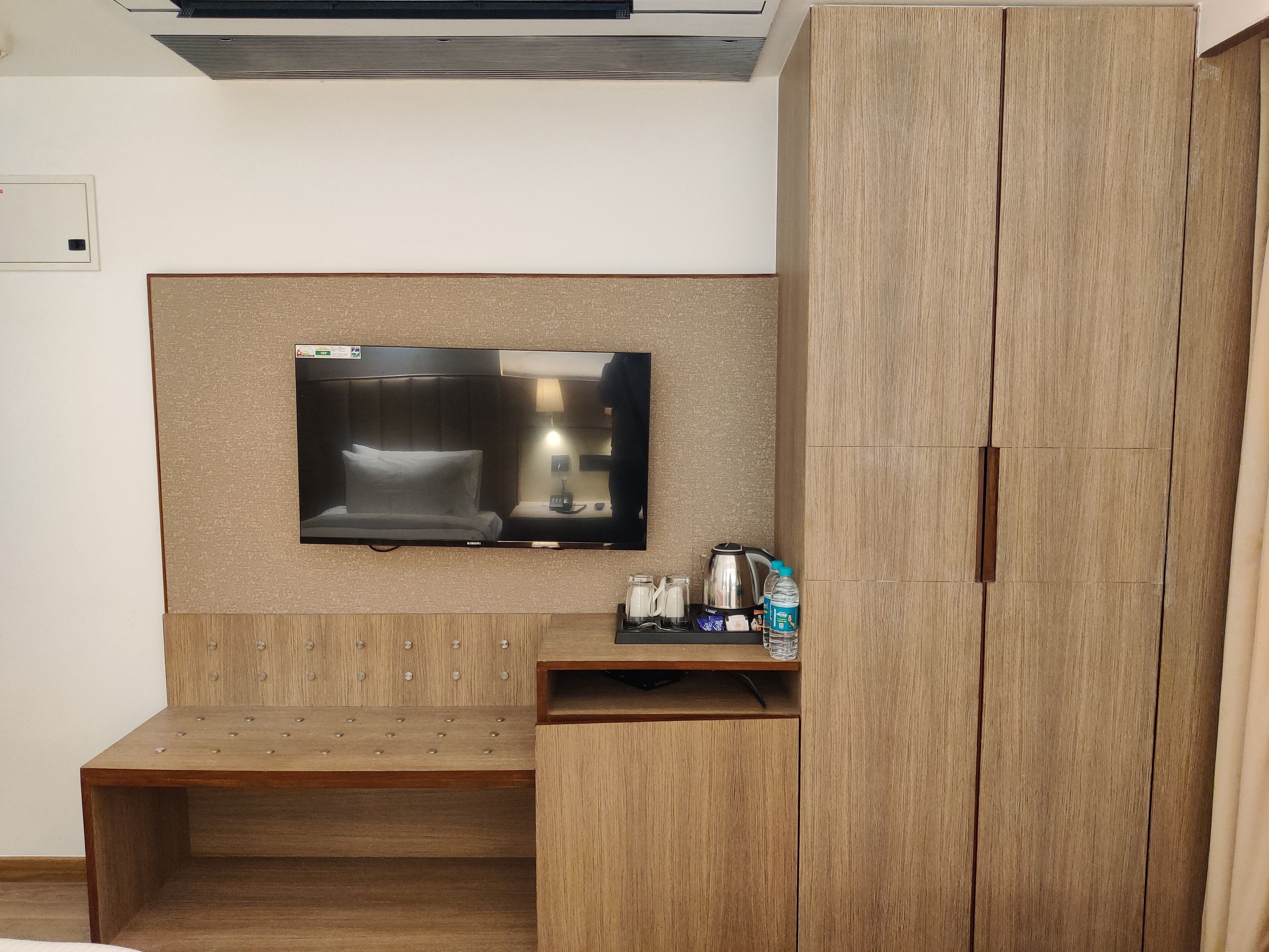 Een hotelkamer met een flatscreen televisie bevestigd aan een gestructureerde muur, een houten bankje, een klein tafeltje met een waterkoker en kopjes, en een kledingkast.