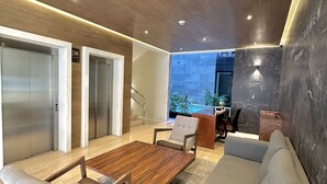 A modern living room with a wooden ceiling, a grey sofa, a wooden coffee table, and two armchairs. There are two elevators in the background. - Modern Pool View New Condo Gym Jacuzzi (Playa del Carmen)