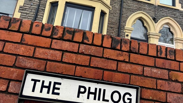 Un edificio con un amplio ventanal en esquina, un muro de ladrillo con un letrero que dice 'THE PHILOG' y fachadas de piedra y pintura.