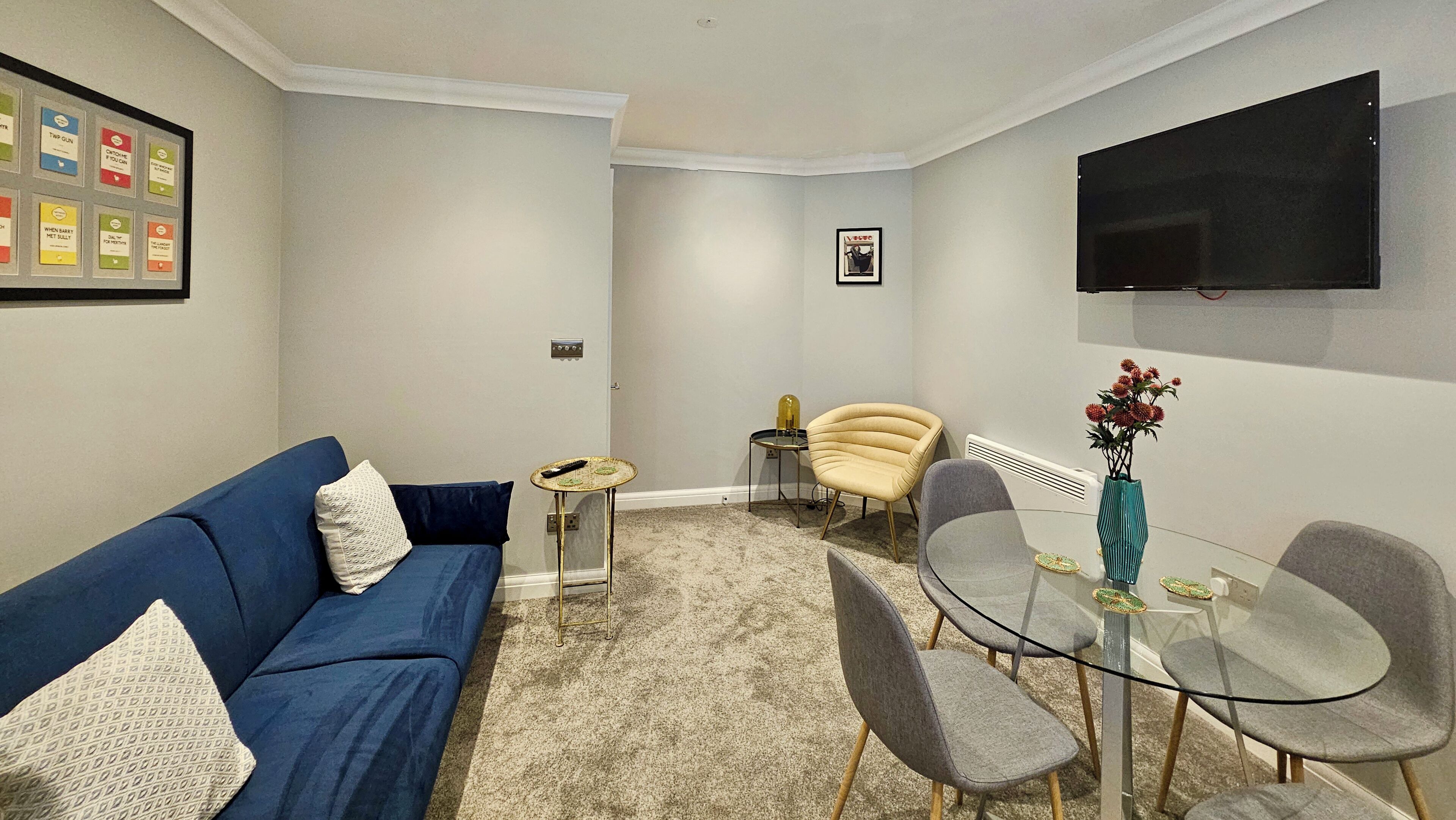 A compact living space with a blue sofa, a glass table with chairs, a small side table, and a mounted TV.