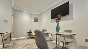 Un salón de conferencias con una mesa de vidrio, sillas tapizadas en gris, un jarrón azul turquesa con flores, un televisor de pantalla plana y suelo alfombrado.