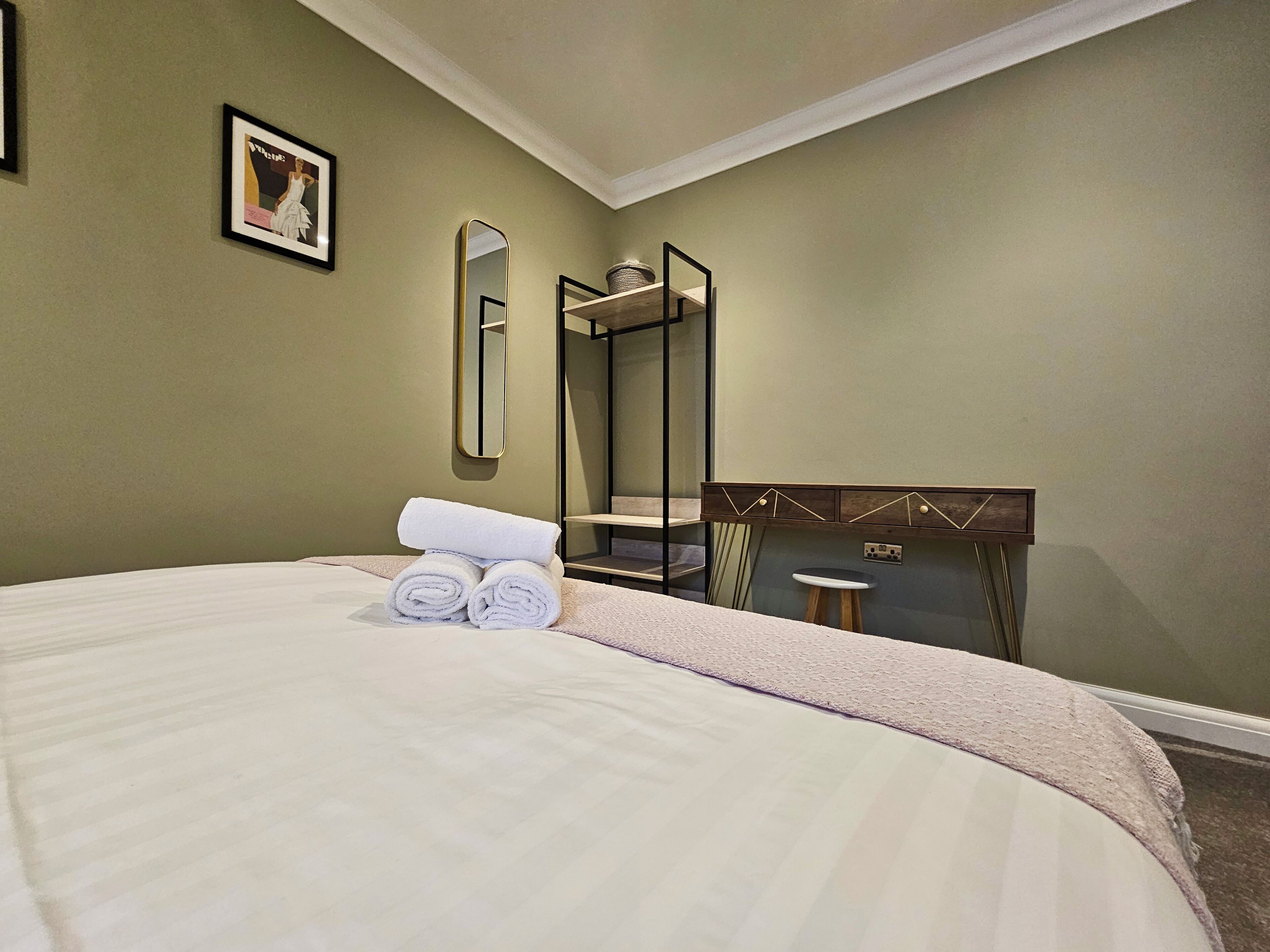 A hotel room with a neatly made bed, a large mirror, a vanity with a stool, and a framed picture on the wall.