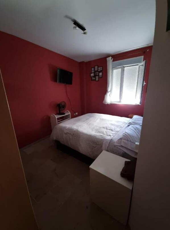 A bedroom with a bed, a desk, a chair, a television on the wall, and a window with blinds. - Orchard Apartment (Lucena)