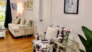 Uma sala de estar com sofá, poltrona e mesa de centro de vidro. Há uma porta e um quadro na parede.
