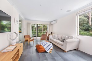 Ein modernes Wohnzimmer mit Sofa, Sesseln, Couchtisch, Fernseher und einem großen Fenster mit Blick auf Grünflächen.
