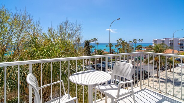 Un balcon avec des chaises et une table en métal blanc, donnant sur une rue bordée de voitures garées et de palmiers, et offrant une vue sur l’océan et les bâtiments au loin.