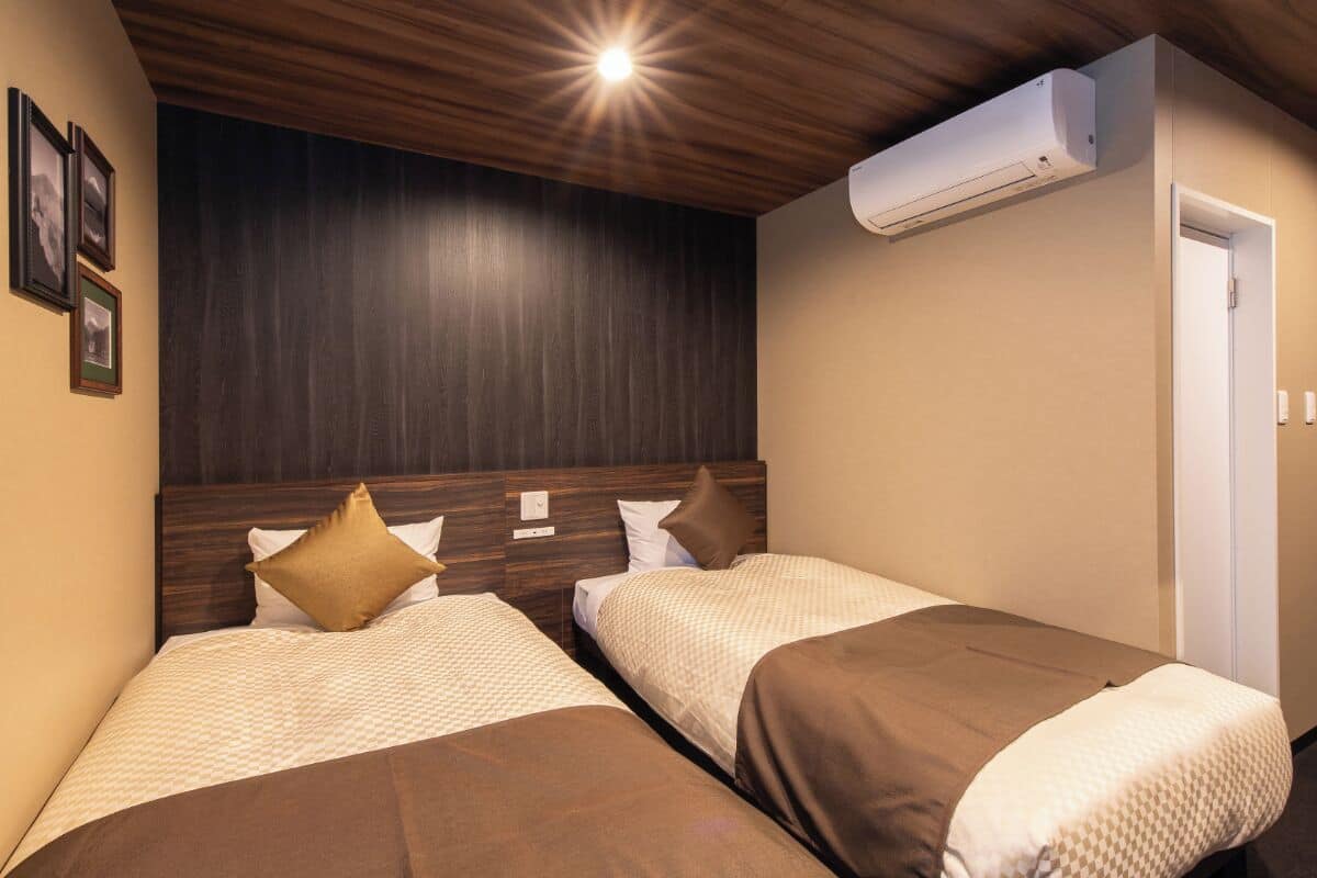 A hotel room with two beds, wooden headboards, a ceiling light, and an air conditioning unit.