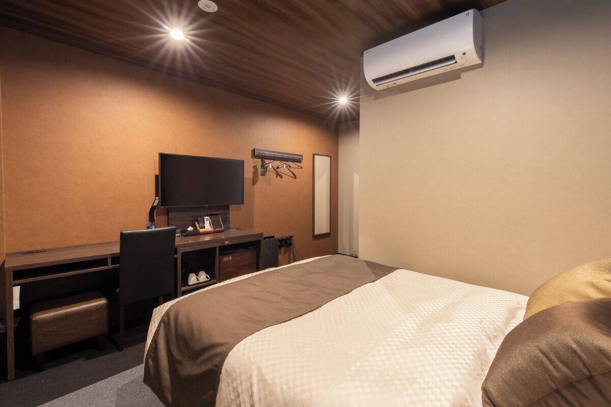 A hotel room with a bed, a desk, a chair, a television, and an air conditioning unit.