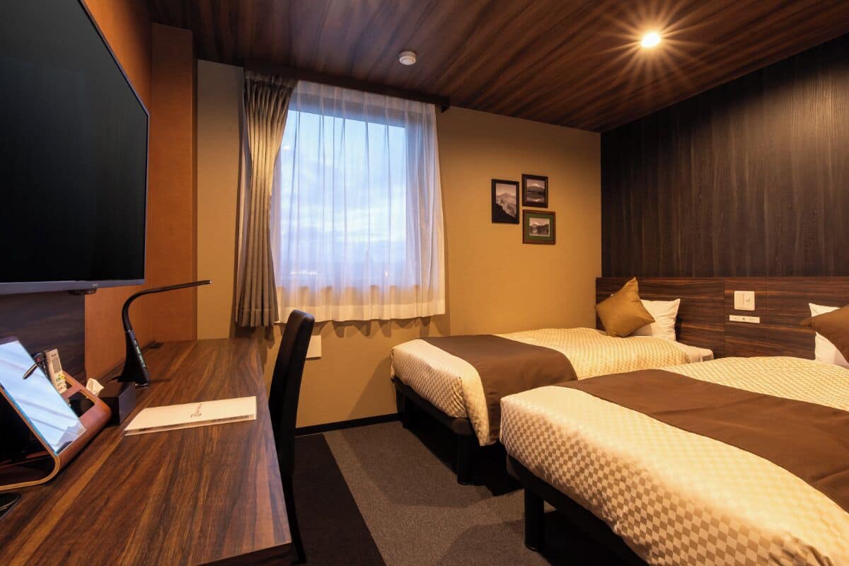 A hotel room with two beds, a desk with a computer, a TV, and a window with curtains.