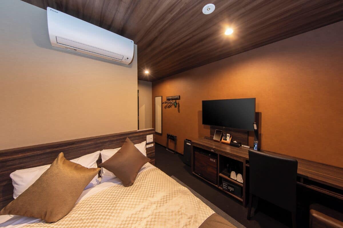 A hotel room with a bed, a TV, a desk, and an air conditioning unit.