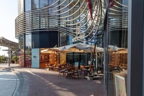 Un bâtiment moderne doté d’une façade en verre incurvé et d’un espace extérieur aménagé avec des parasols.