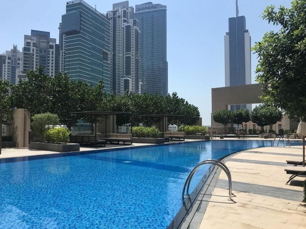 A swimming pool with a metal handrail, surrounded by lounge chairs and potted plants, with high-rise buildings in the background.