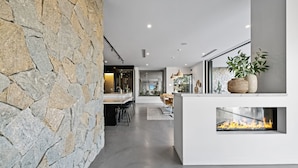 A modern kitchen with a stone wall, a built-in fireplace, and a dining area with bar stools. - Mana Beach House | Luxury Beach House in Kingscliff (Kingscliff)