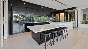 A modern kitchen with a large island, black bar stools, and a view of tropical plants. - Mana Beach House | Luxury Beach House in Kingscliff (Kingscliff)