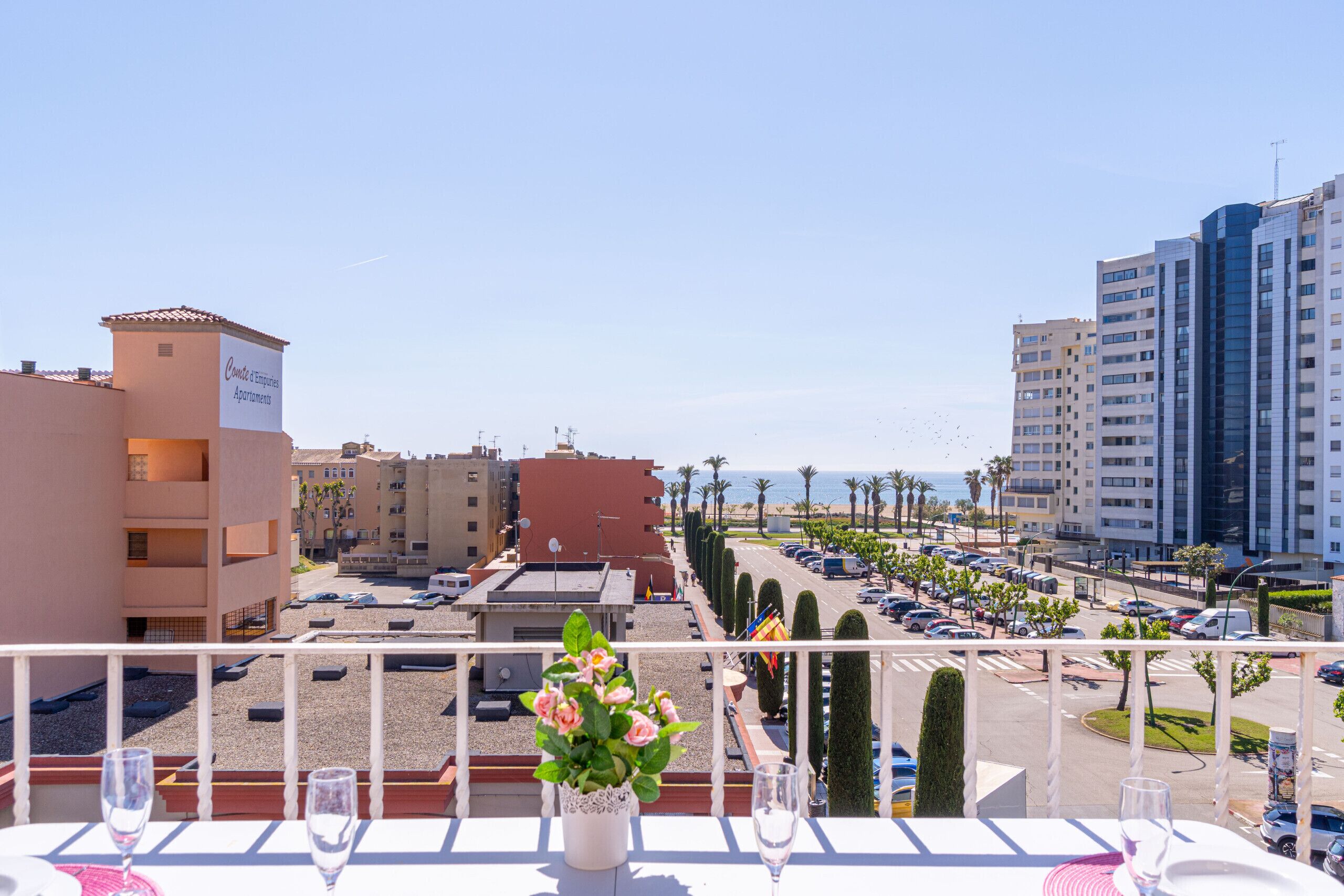 A balcony with a table set for dining, overlooking a parking area and buildings.